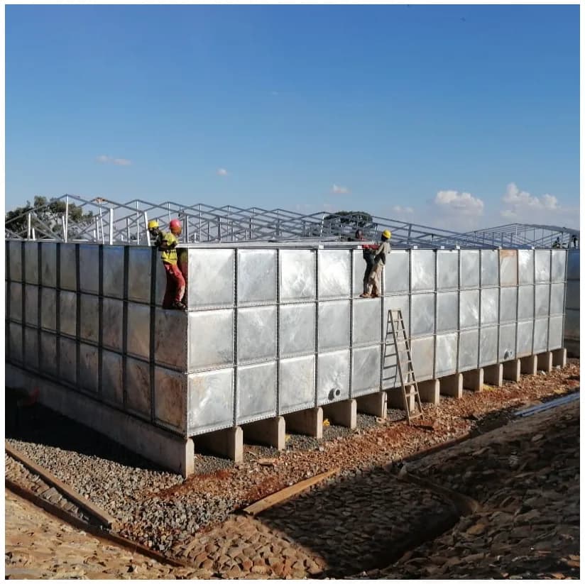 Sectional steel water storage tank under construction on concrete plinths, with steel roof trusses being installed and stone-pitched drainage alongside